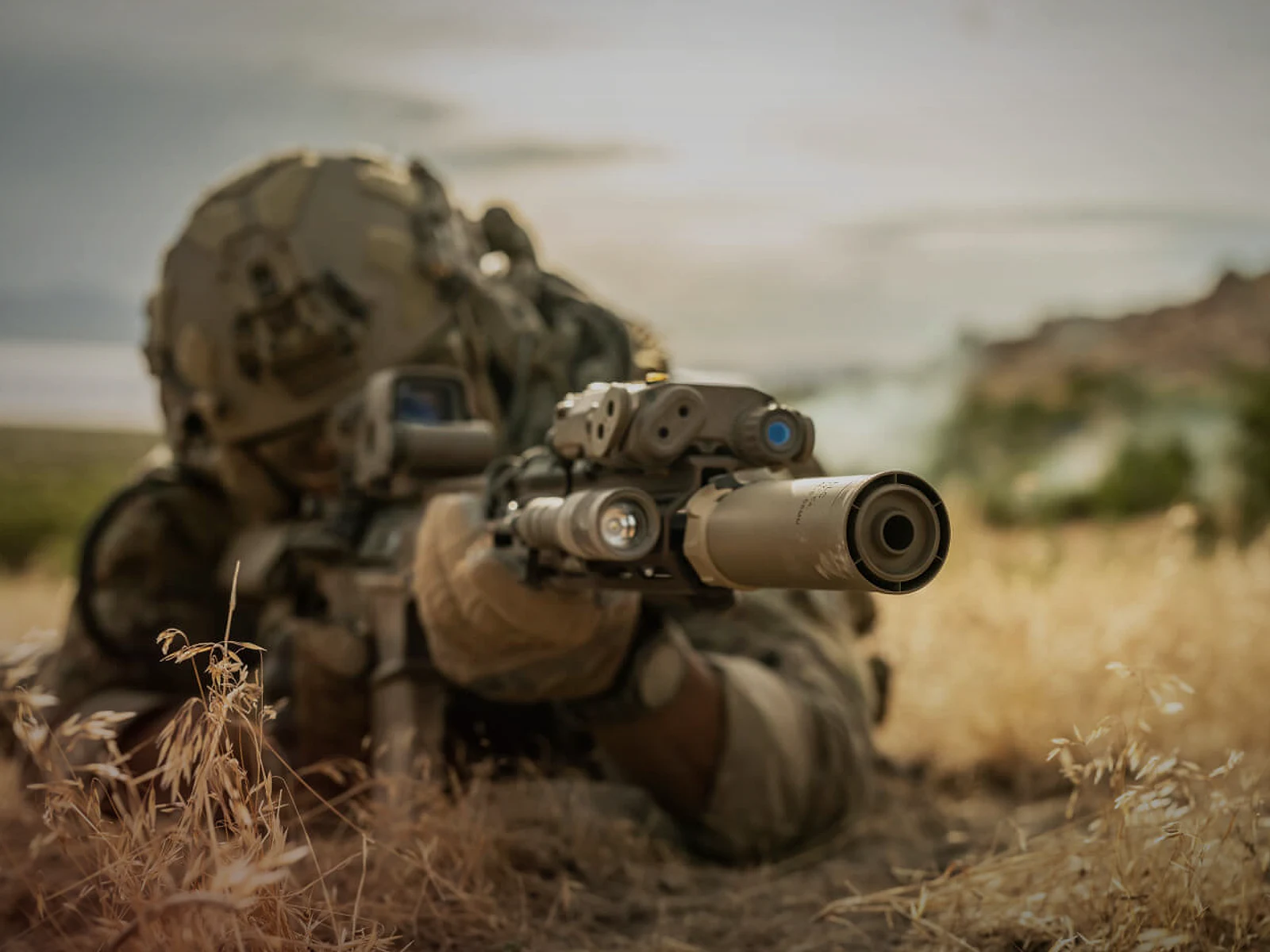 Shooter aiming a suppressed rifle from a prone field position